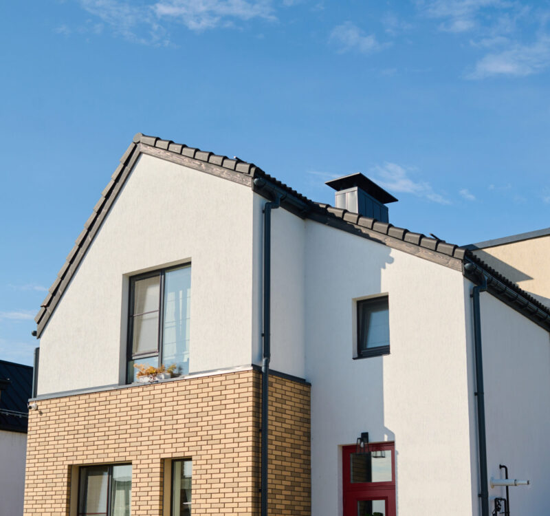 Facade of small white cottage with three windows and closed door standing in front of camera in rural environment