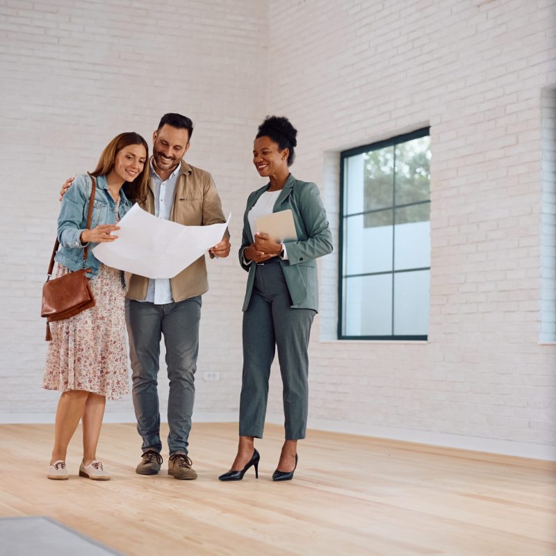 Happy couple of real estate buyers examining blueprints with their agent. Copy space.